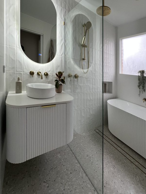 Finished bathroom vanity with wall mounted brass tapware, a round basin and an oval backlit mirror