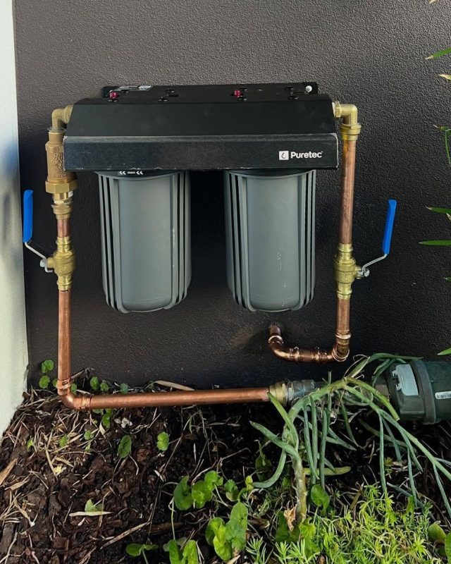 Twin cartridge Puretec water filter plumbed in copper against a dark rendered wall with blue handled isolation valves either side