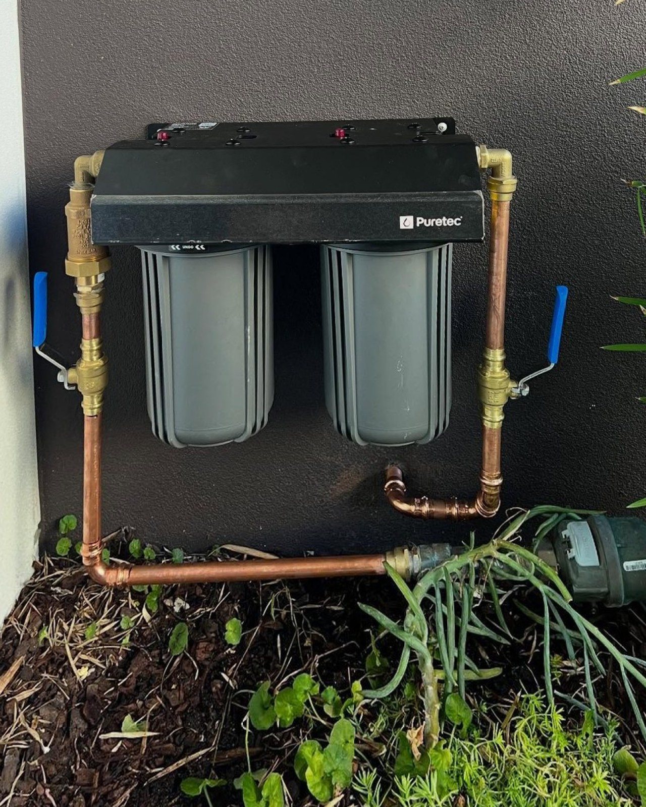 Twin cartridge whole-house water filter plumbed in copper against a dark wall